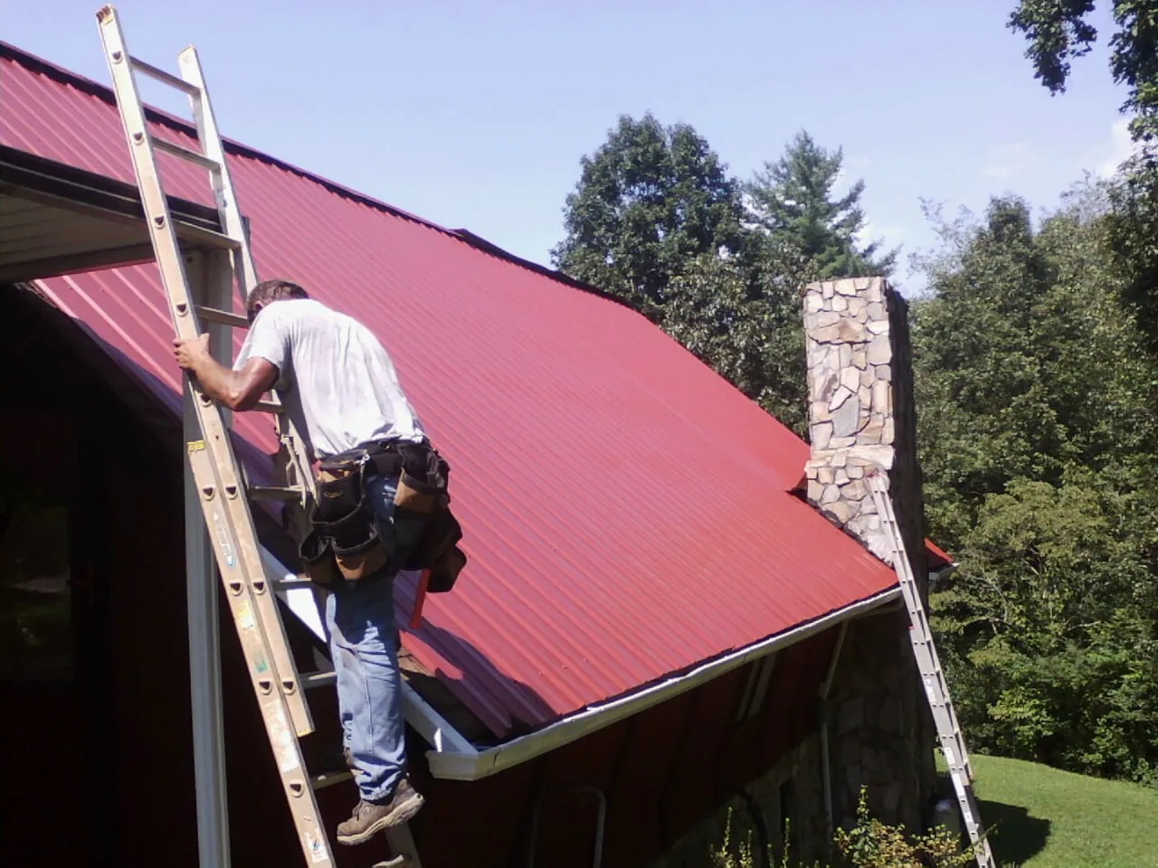 Roofer on a ladder beside a red metal roof and stone chimney — Soffit & Fascia Repair by Roofing Brothers in Cuddebackville